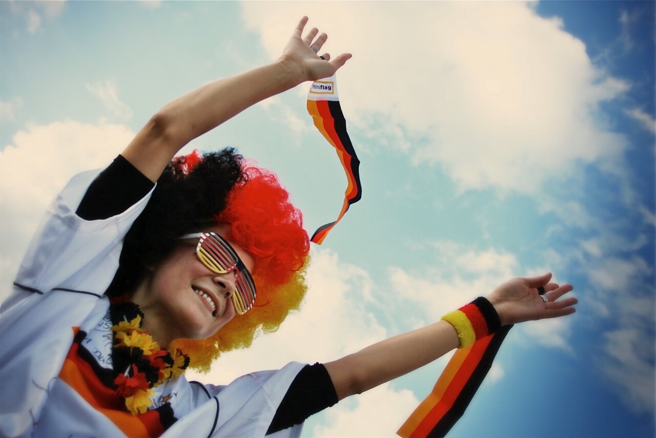 Deutschland Fan in scharz-rot-goldener Fankleidung jubelt. Über ihr sieht man einen hellblauen Himmel.