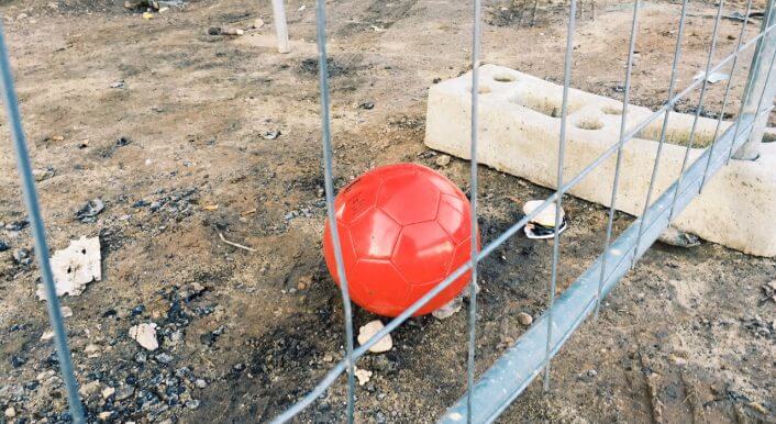 Ein roter Ball liegt hinter einem Bauzaun auf brauner Erde.