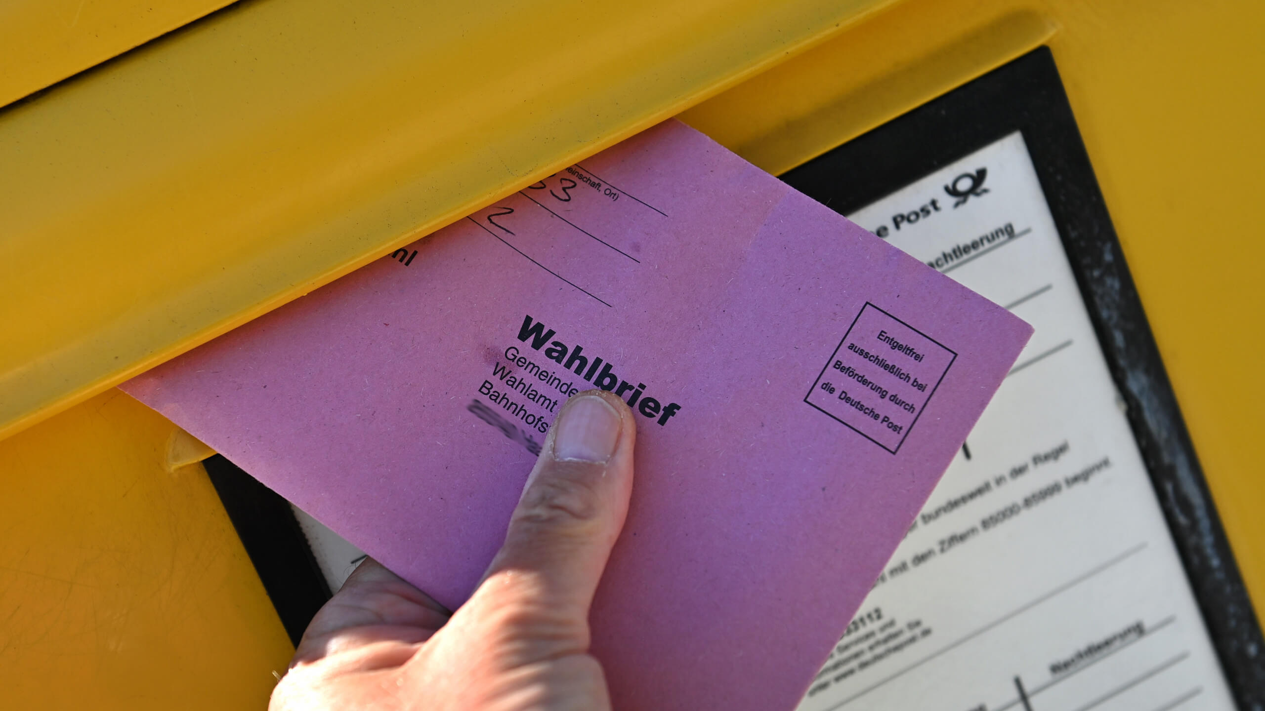 Jemand wirft einen roten Wahlbriefumschlag in einen Briefkasten.