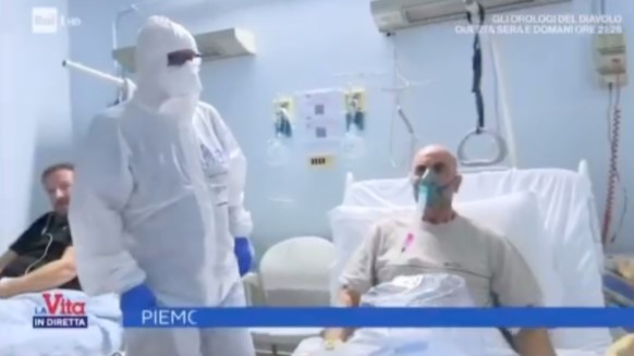 Mann ohne Maske in einem Krankenhausbett, umgeben von Patienten und Pflegern, die alle Masken und teilweise Schutzanzüge tragen.