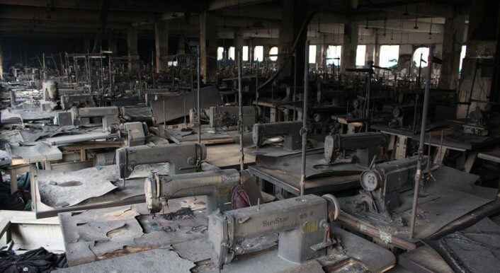 Eine Reihe verkohlter Nähmaschinen steht in einer finsteren Halle.