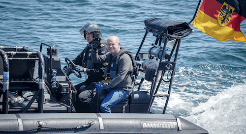 Bundeskanzler Olaf Scholz wird bei seinem Besuch der Deutschen Marine in Rostock-Warnemünde am Montag, 5. Juni 2023, auf einem Boot von der Fregatte "Mecklenburg-Vorpommern" zu einem Minensuchboot gefahren. Das Boot hat den Namen Boomeranger, Scholz lächelt und winkt.