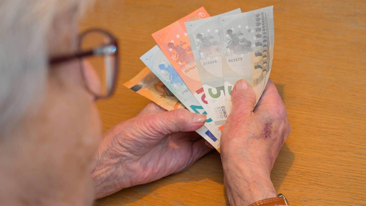 Symbolbild Rente: Frau mit grauen Haaren sitzt am Tisch und hält Geld in der Hand