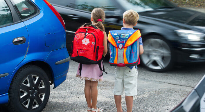Zwei Schulkinder stehen an einer befahrenen Straße, die sie überqueren wollen