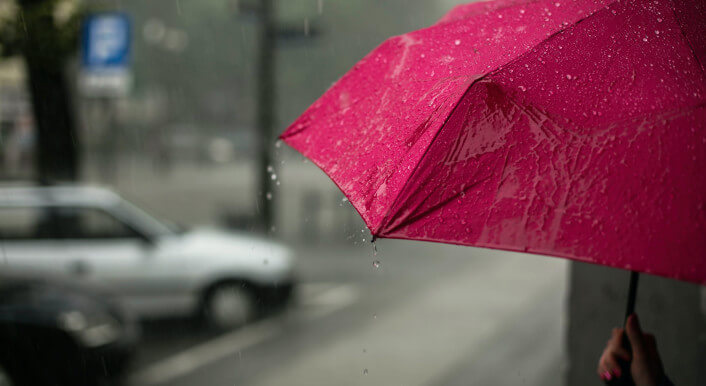 Eine Hand hält einen roten Regenschirm von rechts in das Bild hinein. Im Hintergrund ist verschwommen die Szenerie einer Straße zu erkennen.
