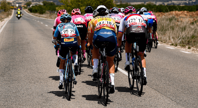 Mehrere Menschen in Sportkleidung fahren auf Fahrrädern eine Straße während La Vuelta entlang.