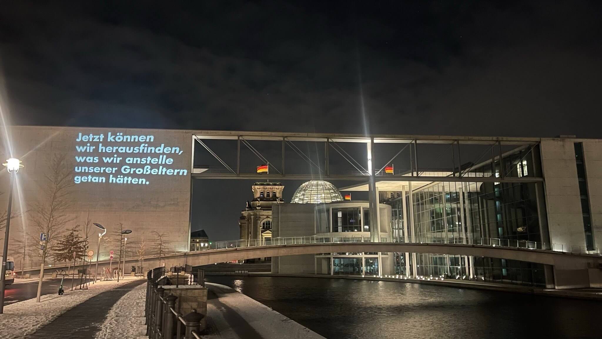 Nacht in Berlin: Im Hintergrund ist der Reichstag zu sehen, davor der Bundestag mit einer Projektion mit dem Satz: „Jetzt können wir herausfinden, was wir anstelle unserer Großeltern getan hätten“