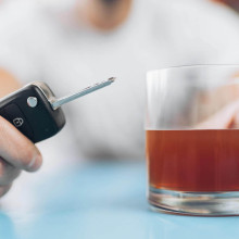 Eine Person an einem Tisch, verschwommen, sie hält einen Autoschlüssel und ein alkoholisches Getränk im Glas in der Hand