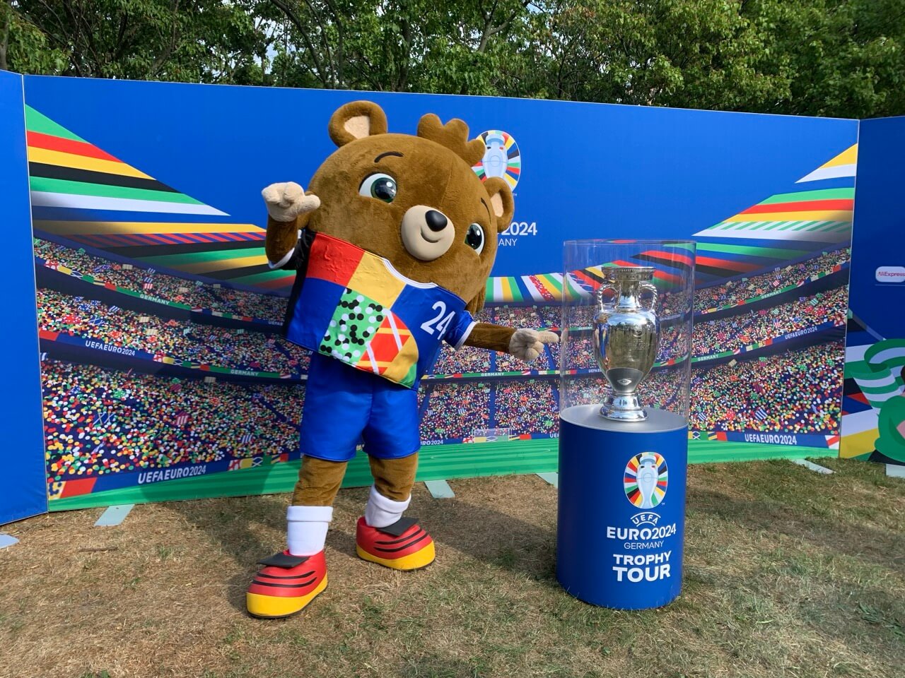 Links: Der Teddybär Älbärt im bunten Trikot, das Maskottchen für die Europameisterschaft 2024 in Deutschland. Rechts eine Nachbildung des EM-Pokals.