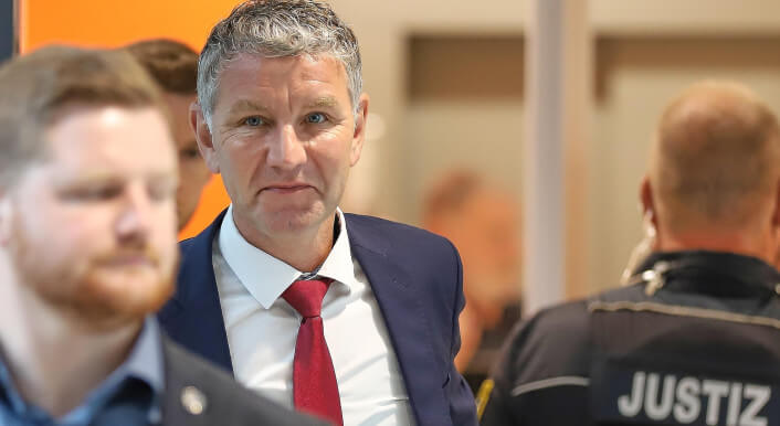 Der AfD-Politiker ist im Fokus des Bilds, er trägt Anzug und Krawatte. Höcke steht im Amtsgericht Halle. Vor ihm läuft ein weiterer Mann, neben ihm steht eine Person in Uniform mit der Aufschrift „Justiz".