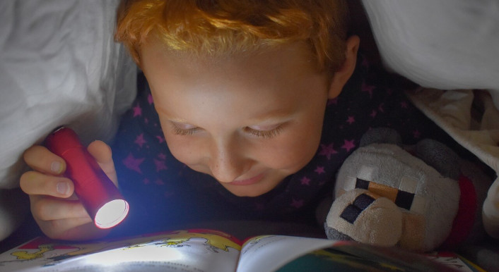 Kind liest unter der Bettdecke mit einer Taschenlampe.