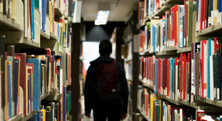 Ein Kind mit Schulranzen steht zwischen Bücherregalen in einer Bibliothek. Foto: redd-f-9o8Y / unsplash.com