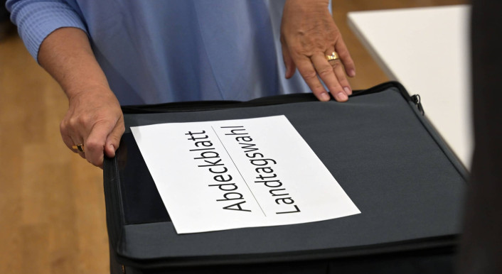 Mann legt ein A4-Blatt mit der Aufschrift Abdeckblatt Landtagswahl auf eine Wahlurne aus Stoff mit Reißverschluss
