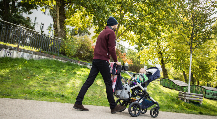 Mann mit Kleinkind schiebt einen Kinderwagen durch einen Park