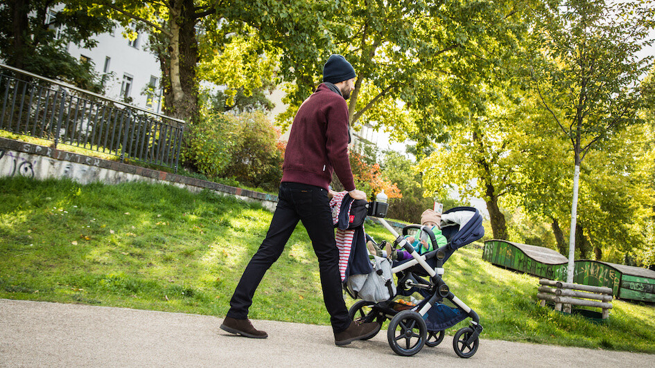 Mann mit Kleinkind schiebt einen Kinderwagen durch einen Park