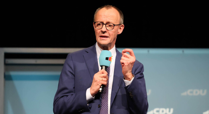 Friedrich Merz im Anzug vor einer blauen Wand mit dem CDU-Logo – mit Mikrofon in der Hand