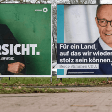 Wahlplakate von Robert Habeck und Friedrich Merz für die Bundestagswahl an einer Straße – ein Radfahrer fährt vorbei.