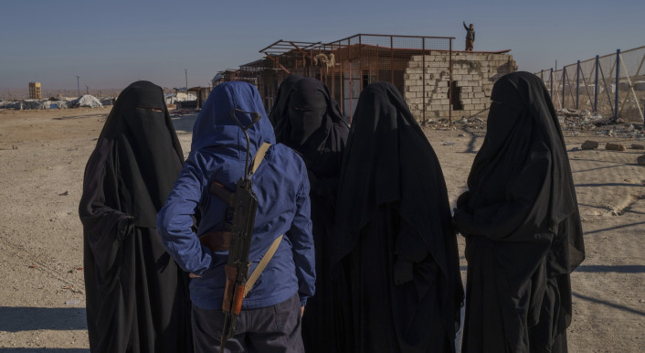 Eine Gruppe schwarz verschleierter Frauen steht in einem Brachland und spricht mit einem Mann in blauem Anorak. Im Hintergrund sieht man Baracken und Zäune.