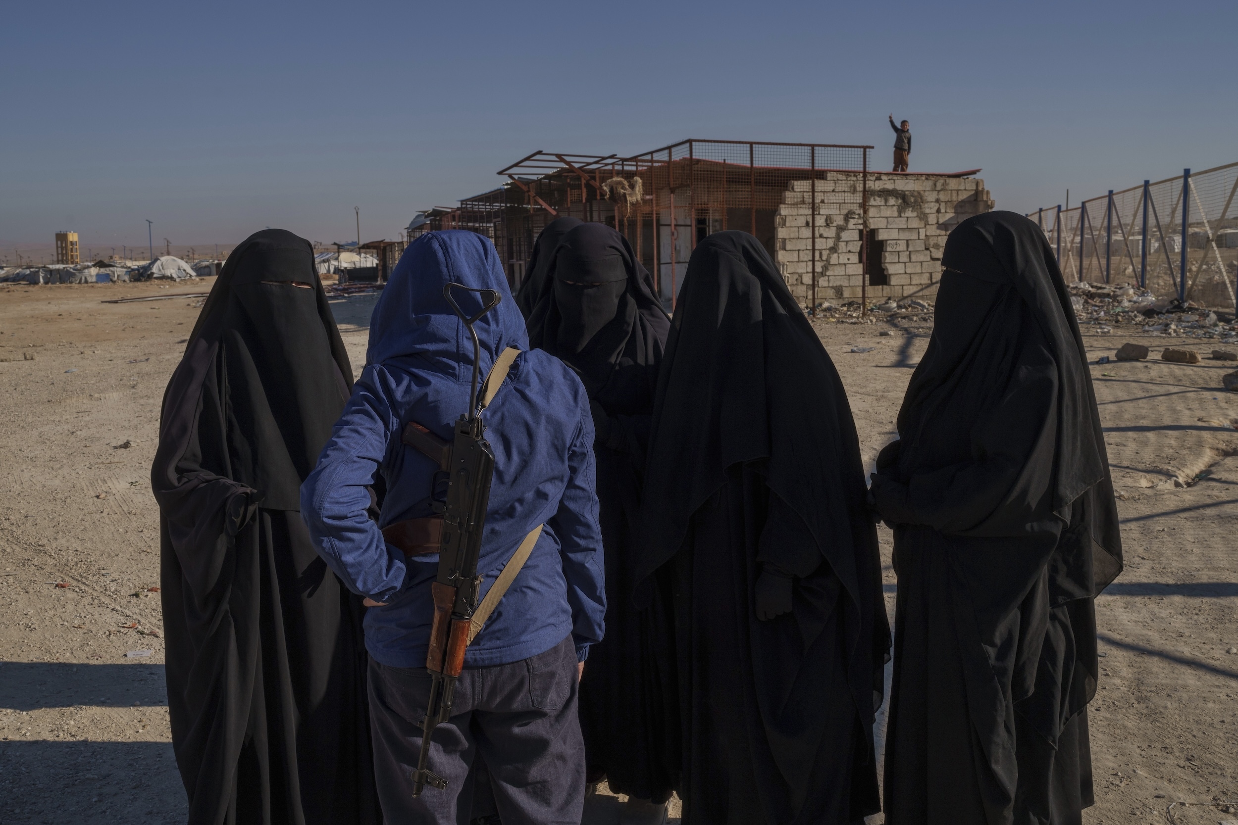 Eine Gruppe schwarz verschleierter Frauen steht in einem Brachland und spricht mit einem Mann in blauem Anorak. Im Hintergrund sieht man Baracken und Zäune.