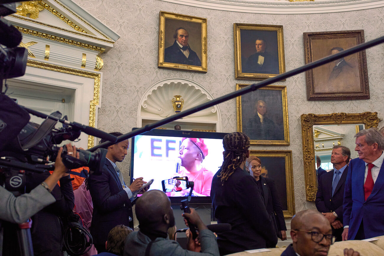 Screen mit Video im Weißen Haus, davor mehrere Pressevertreter und andere Personen