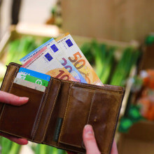 Eine Person hält ein Lederportemonnaie, aus dem mehrere Geldscheine herausstehen. Im Hintergrund ist eine GGemüseauslage zu sehen.