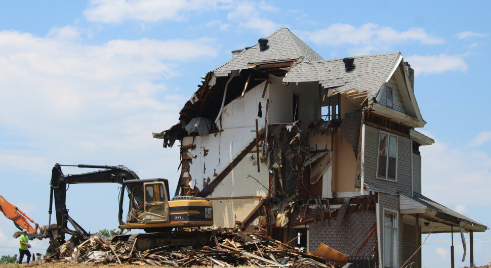 An excavator in front of a half-demolished house