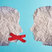 Zwei ausgeschnittene Gesichter aus Papier stehen sich gegenüber, eines hat ein rotes X vor dem Mund.