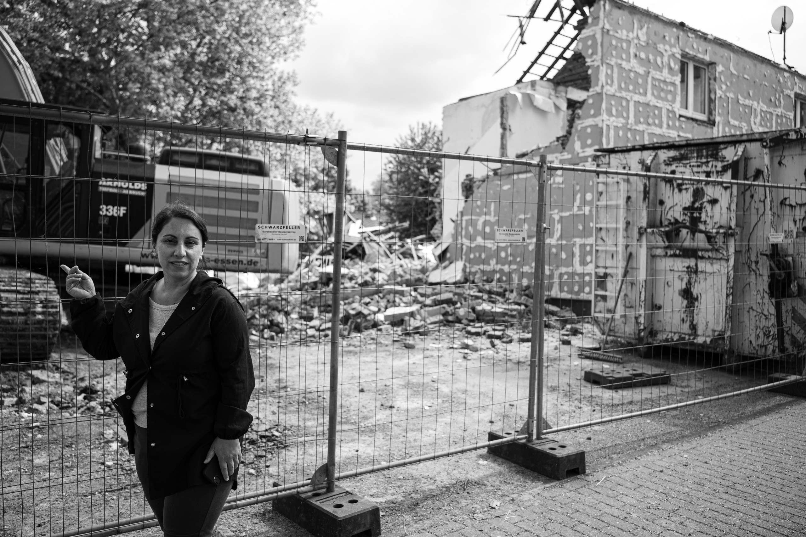 Eine Frau steht vor einem Bauzaun, dahinter steht ein Bagger, der ein Gebäude bereits zum Teil abgerissen hat.