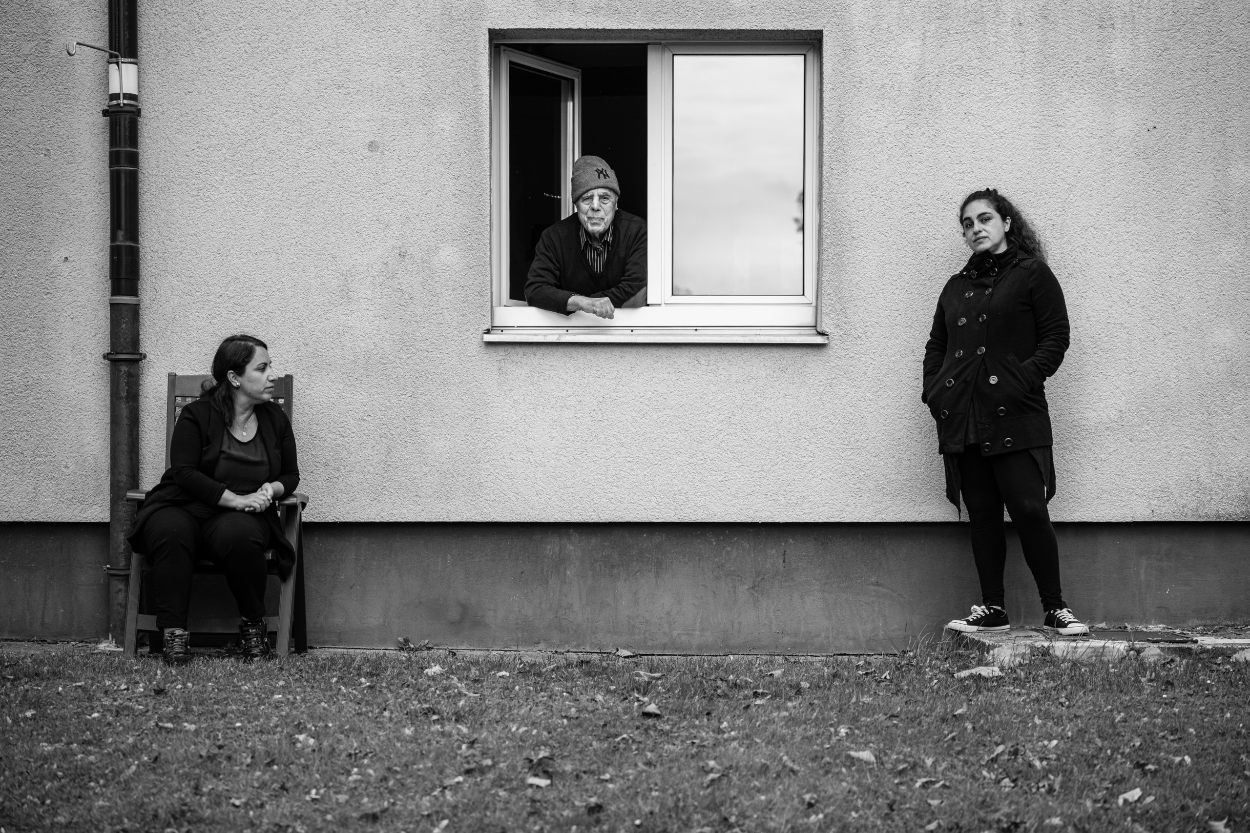 Zwei Personen vor einer Hauswand, eine sitzt, eine steht. Eine dritte Person schaut mittag zwischen den beiden anderen Personen aus dem Fenster.