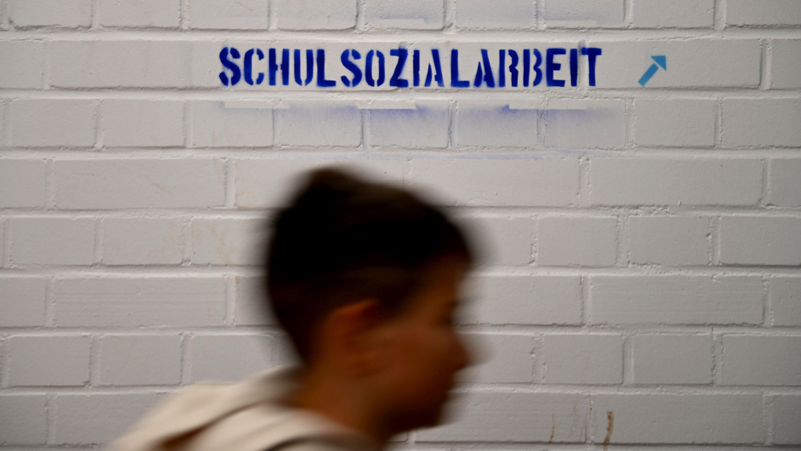 Ein Kind läuft an einer weißen Wand mit dem blauen Schriftzug Schulsozialarbeit vorbei.