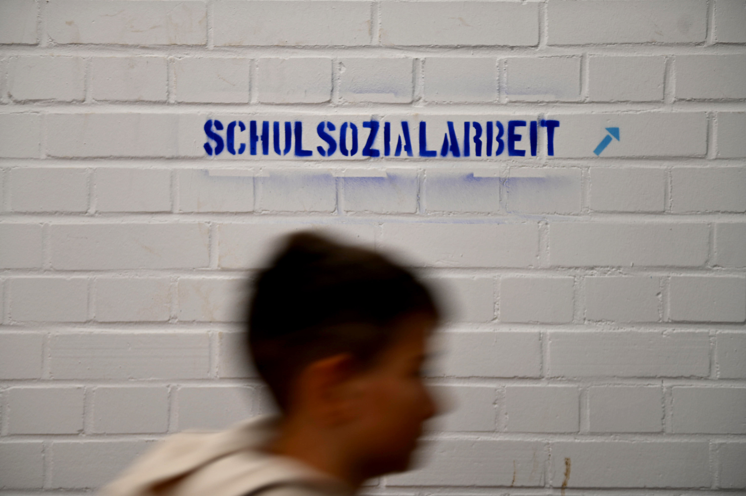 Ein Kind läuft an einer weißen Wand mit dem blauen Schriftzug Schulsozialarbeit vorbei.