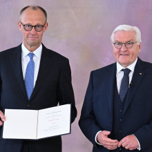 Friedrich Merz steht mit einer Urkunde in der Hand neben Bundespräsident Frank-Walter Steinmeier. Im Hintergrund die Deutschland-Flagge