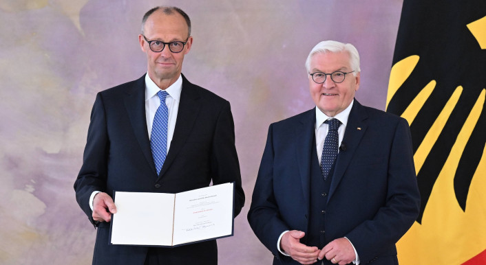 Friedrich Merz steht mit einer Urkunde in der Hand neben Bundespräsident Frank-Walter Steinmeier. Im Hintergrund die Deutschland-Flagge