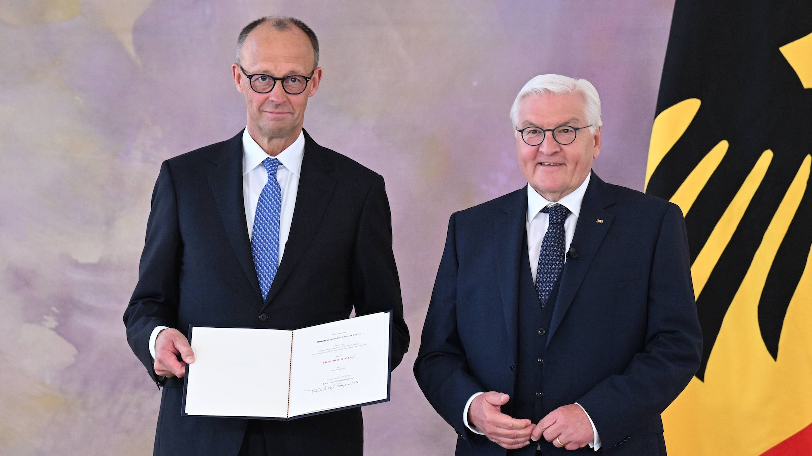 Friedrich Merz steht mit einer Urkunde in der Hand neben Bundespräsident Frank-Walter Steinmeier. Im Hintergrund die Deutschland-Flagge