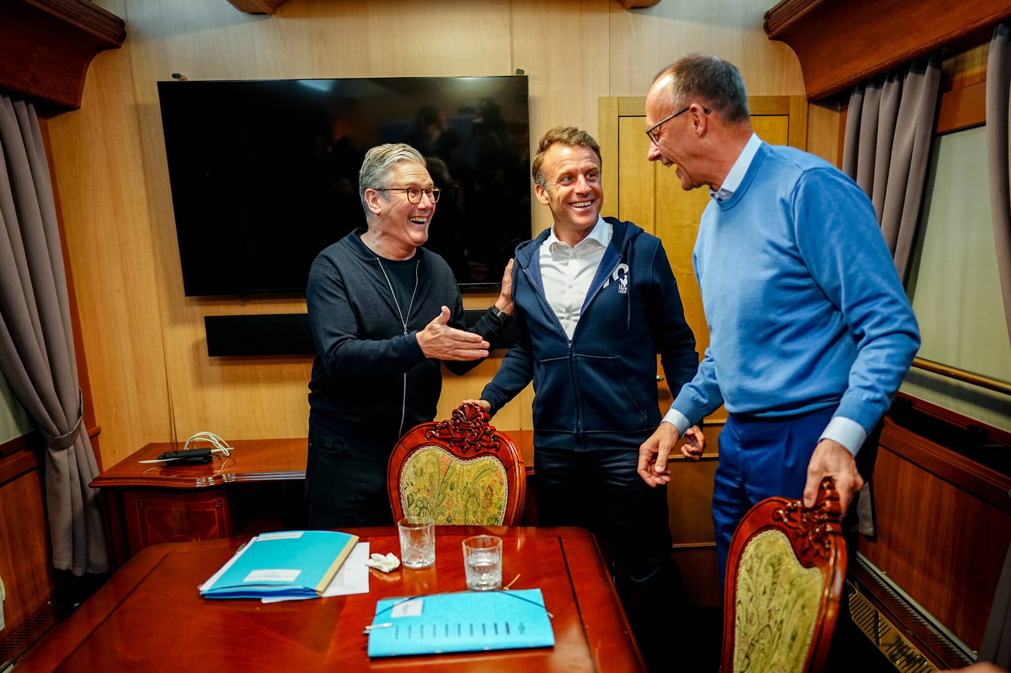 Keir Starmer, Friedrich Merz und Emmanuel Macron begrüßen sich lachend in einem Zug-Abteil. Auf dem Tisch liegen zwei Ordner, 2 Gläser und ein Taschentuch
