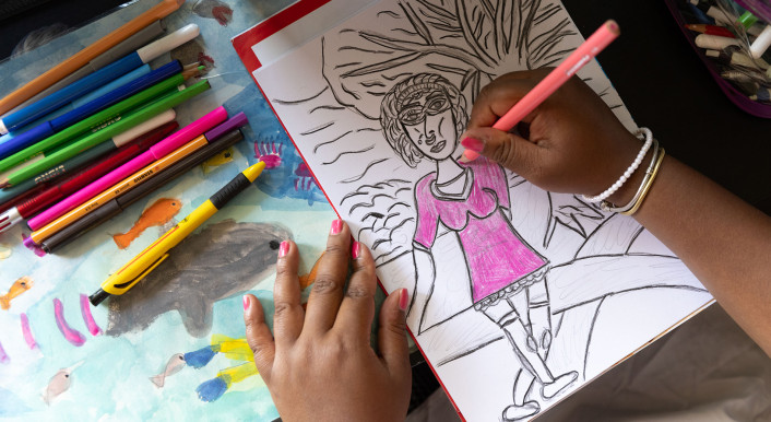 Anonymisiertes Bild der Protagonistin Danah Ayob. Mit Buntstiften malt sie eine Frau im pinken Kleid auf weisses Papier.