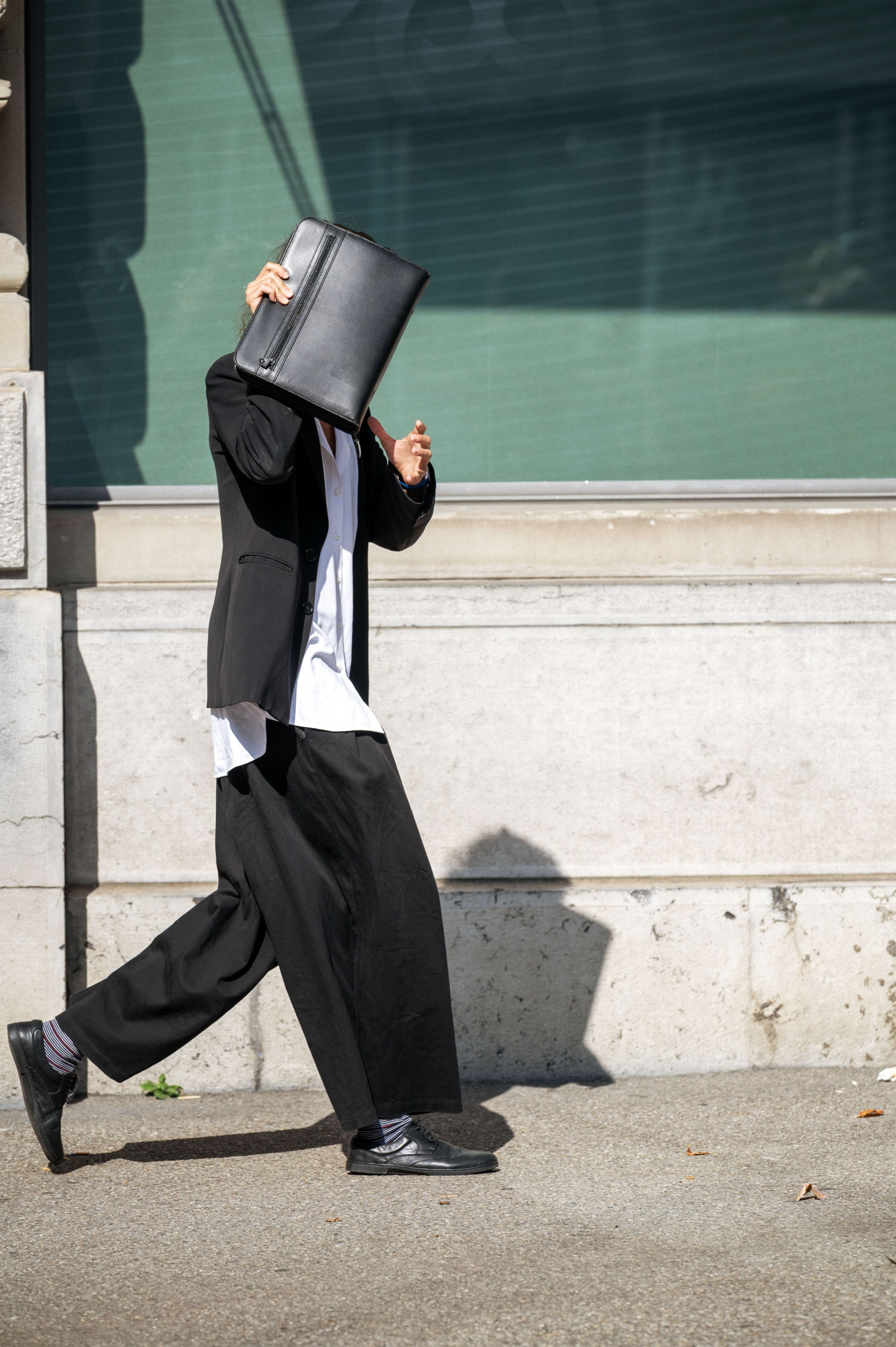 Ein Mann auf der Strasse hält sich eine schwarze Mappe vor das Gesicht. Er will nicht erkannt werden. Er trägt ein weisses Hemd, einen schwarzen Blazer und schwarze weite Hosen.