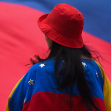 Eine Frau mit rotem Hut von hinten mit ungehangener Venezuela-Flagge, im Hintergrund eine weitere große Venezuela-Flagge