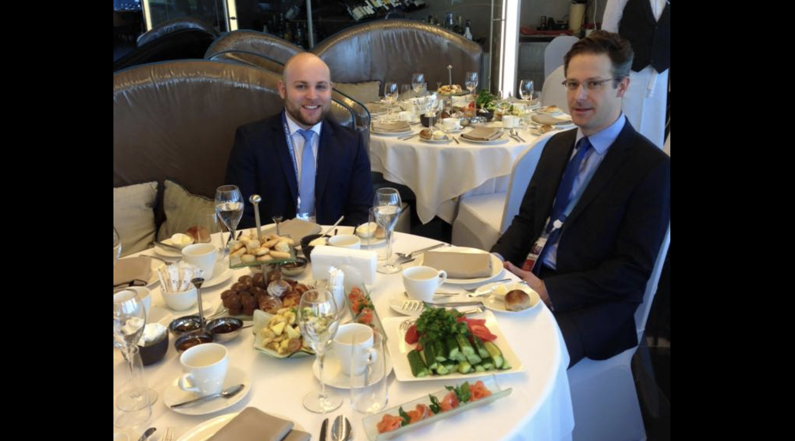 Markus Frohnmaier und Marcus Pretzell sitzen zusammen an einem Tisch voller Speisen und lächeln in die Kamera.