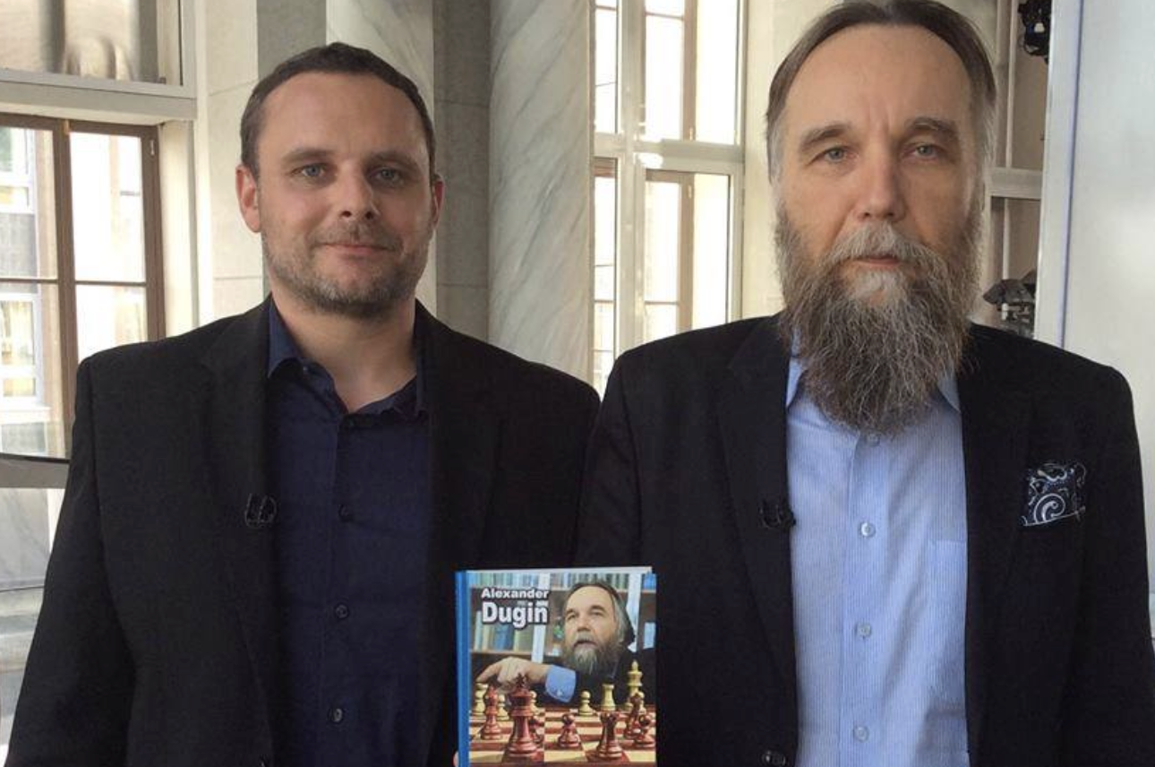 Manuel Ochsenreiter und Alexander Dugin posieren vor einem Buch, auf dem Dugin mit einem Schachbrett abgebildet ist.