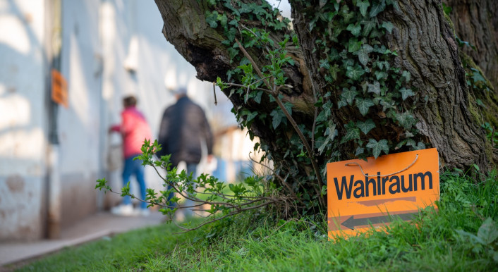 Wahlen in Deutschland: Schild mit der Aufschrift Wahlraum steht vor einem Baum - im Hintergrund sind Menschen und ein Gebäude zu sehen