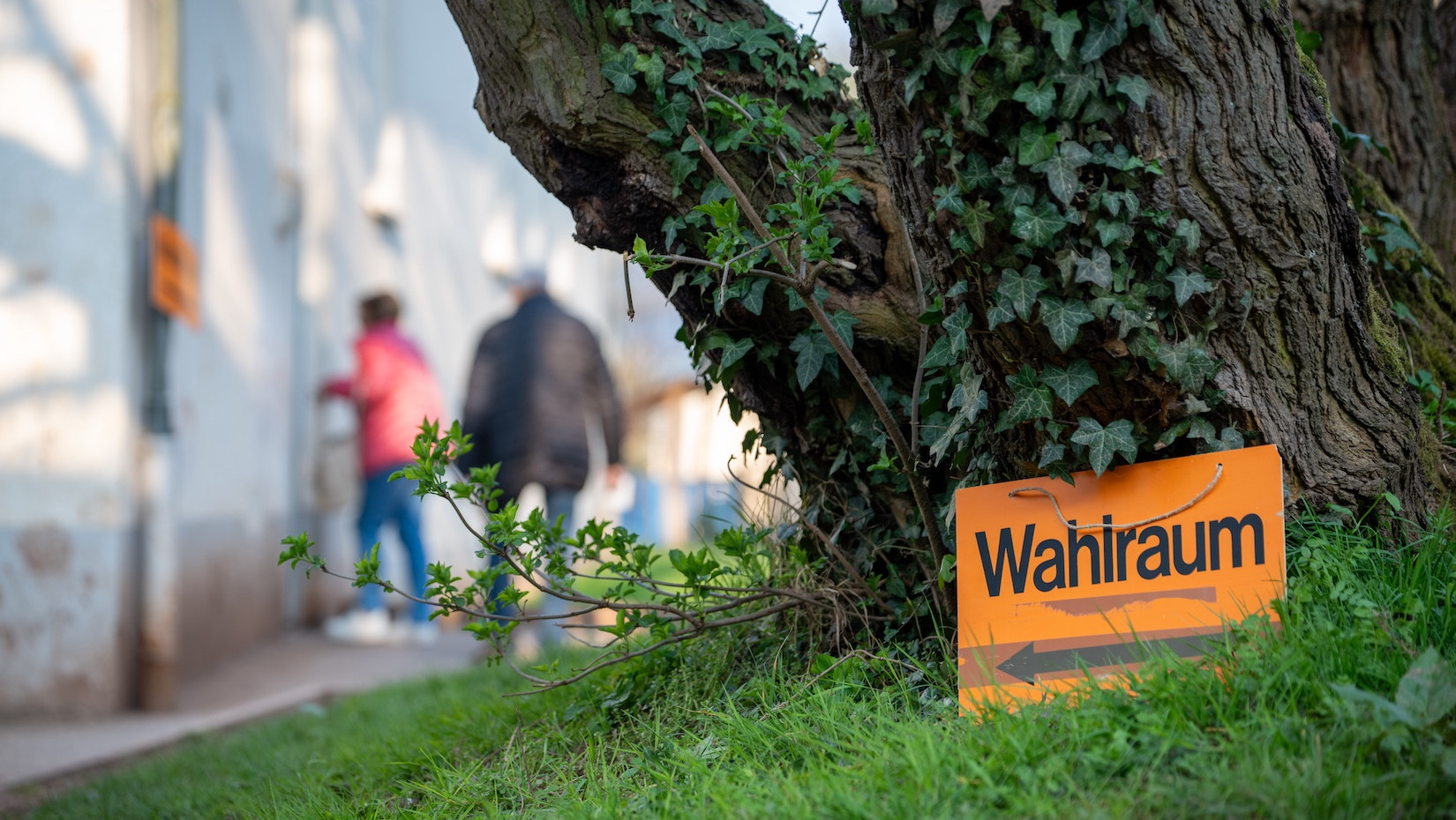Wahlen in Deutschland: Schild mit der Aufschrift Wahlraum steht vor einem Baum - im Hintergrund sind Menschen und ein Gebäude zu sehen