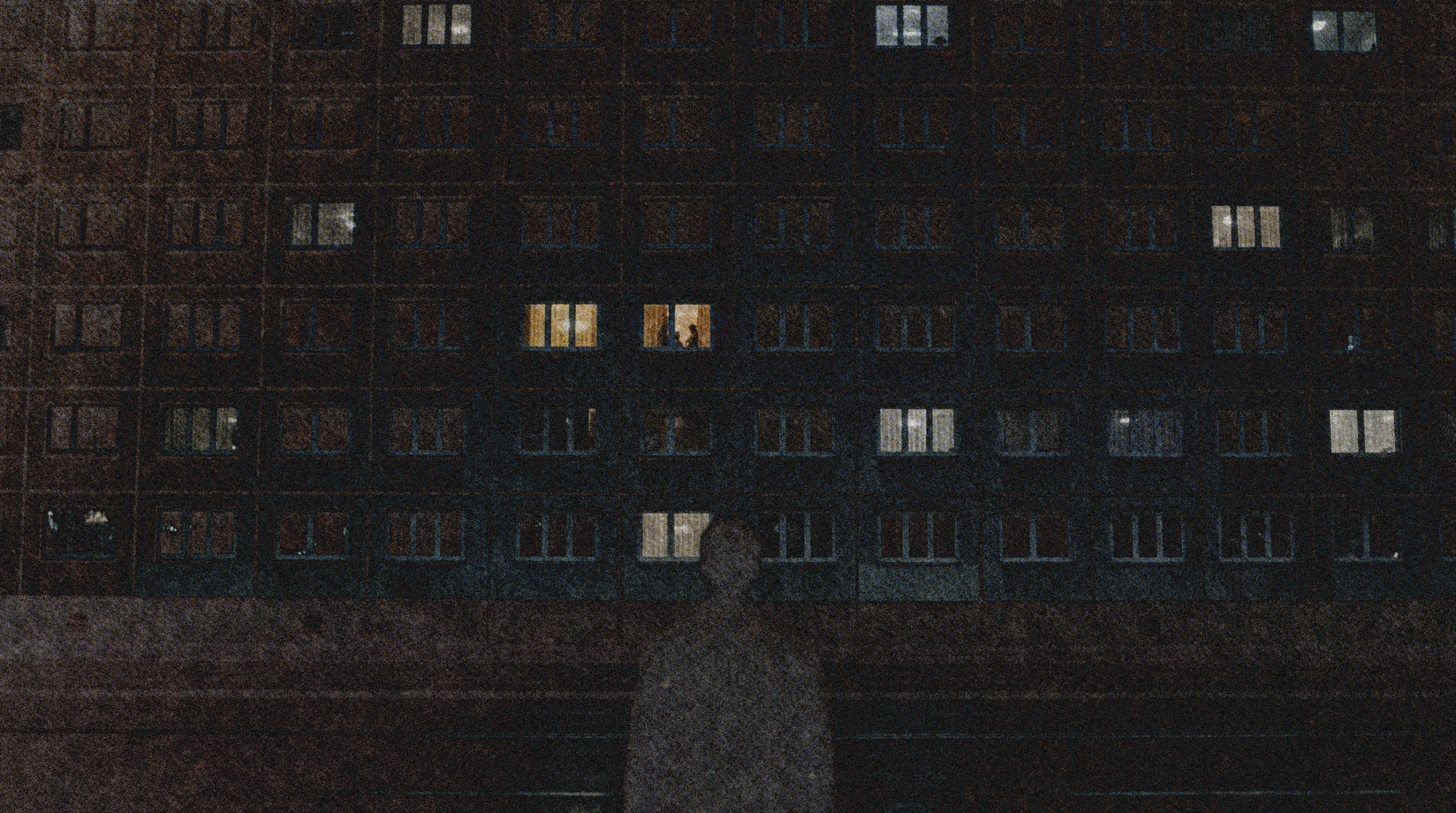 Das Bild zeigt ein Haus bei Nacht, in dem noch wenige Fenster beleuchtet sind. Davor steht eine dunkle Gestalt und beobachtet zwei Personen, die im Haus vor dem Fenster stehen.