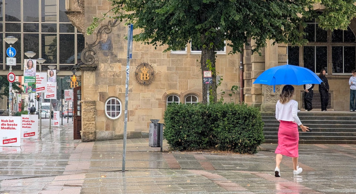 Frau läuft mit Regenschirm über die Straße in Bielefeld, im Hintergrund sind Wahl-Plakate aufgestellt