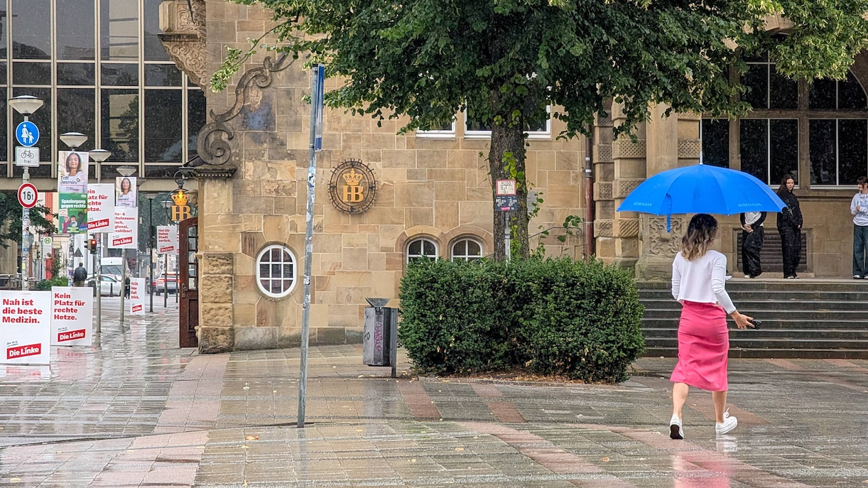 Frau läuft mit Regenschirm über die Straße in Bielefeld, im Hintergrund sind Wahl-Plakate aufgestellt