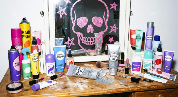 Zu sehen ist ein Schreibtisch mit einem Schminkspiegel. Auf dem Spiegel ist mit pinker Farbe ein Totenkopf gemalt. Auf dem Tisch liegen silberne Tuben, auf denen Glitzergel steht.