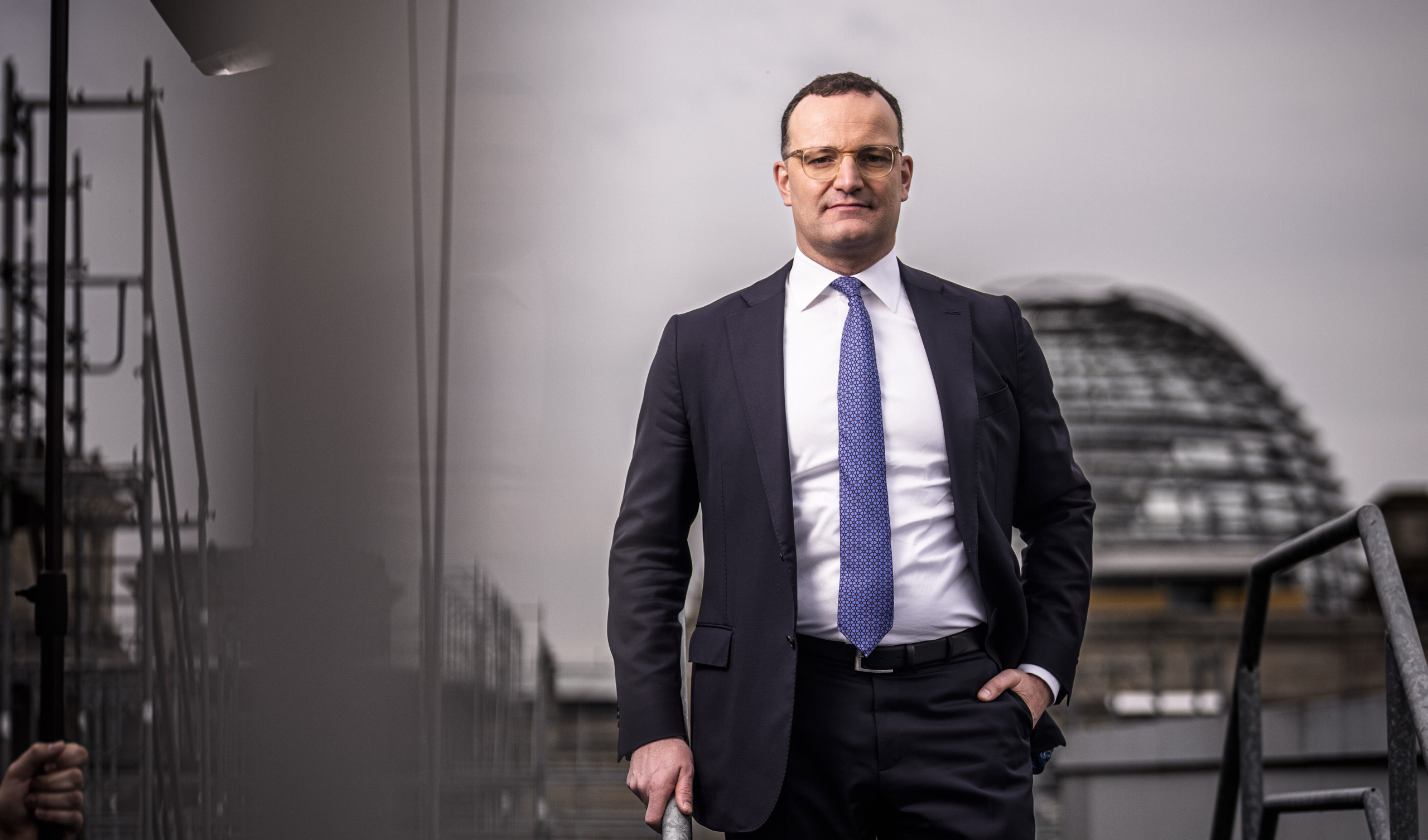 Der Unions-Fraktionsvorsitzende Jens Spahn (CDU) steht an einem Metall-Geländer, er trägt einen schwarzen Anzug. Im Hintergrund ist die Kuppel des Bundestags zu sehen, die Stimmung des Bilds ist düster.