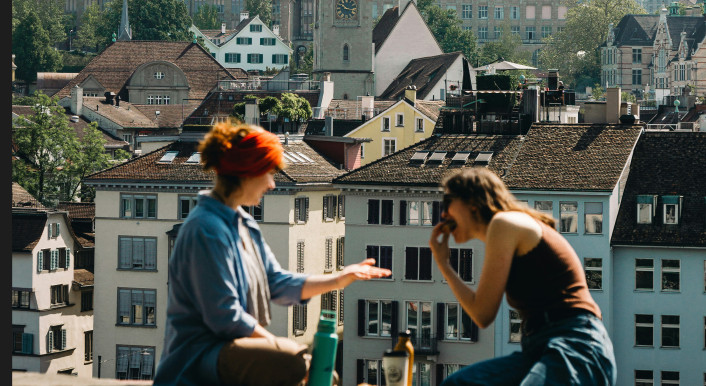 Zwei Frauen sitzen auf einer Mauer vor einem Stadtpanorama in Zürich und essen, reden, trinken zusammen.