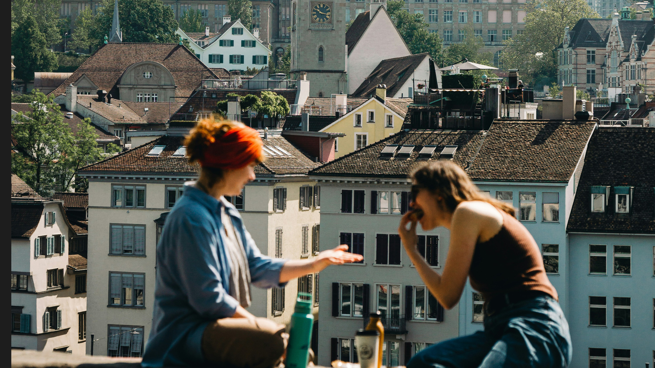 Zwei Frauen sitzen auf einer Mauer vor einem Stadtpanorama in Zürich und essen, reden, trinken zusammen.