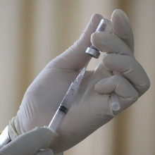 Hand hält aufgezogene Spritze, an deren Kanüle sich ein Tropfen bildet